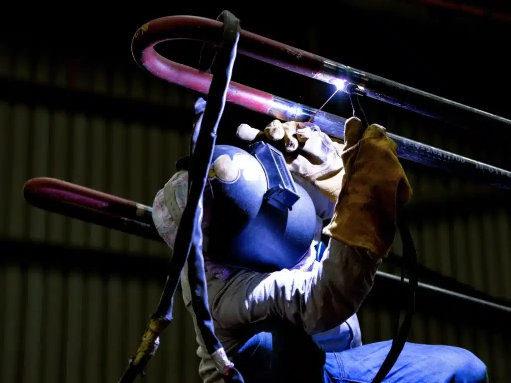 A welder wearing a protective helmet and gloves performs arc welding on overhead metal pipes in a dark industrial workshop.