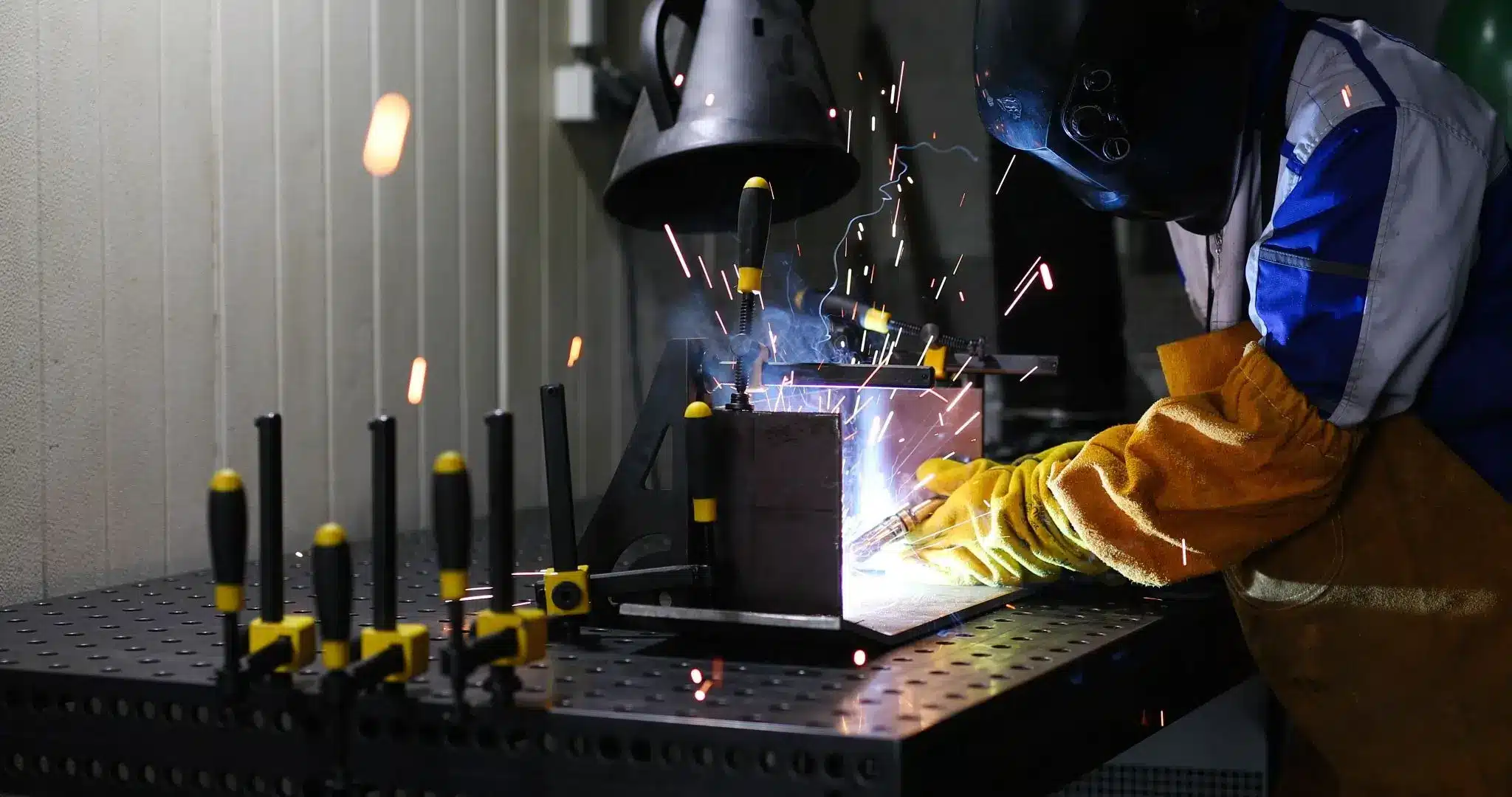 A welder in protective helmet and gloves welds a clamped metal workpiece on an industrial table, with bright sparks and smoke in a workshop.