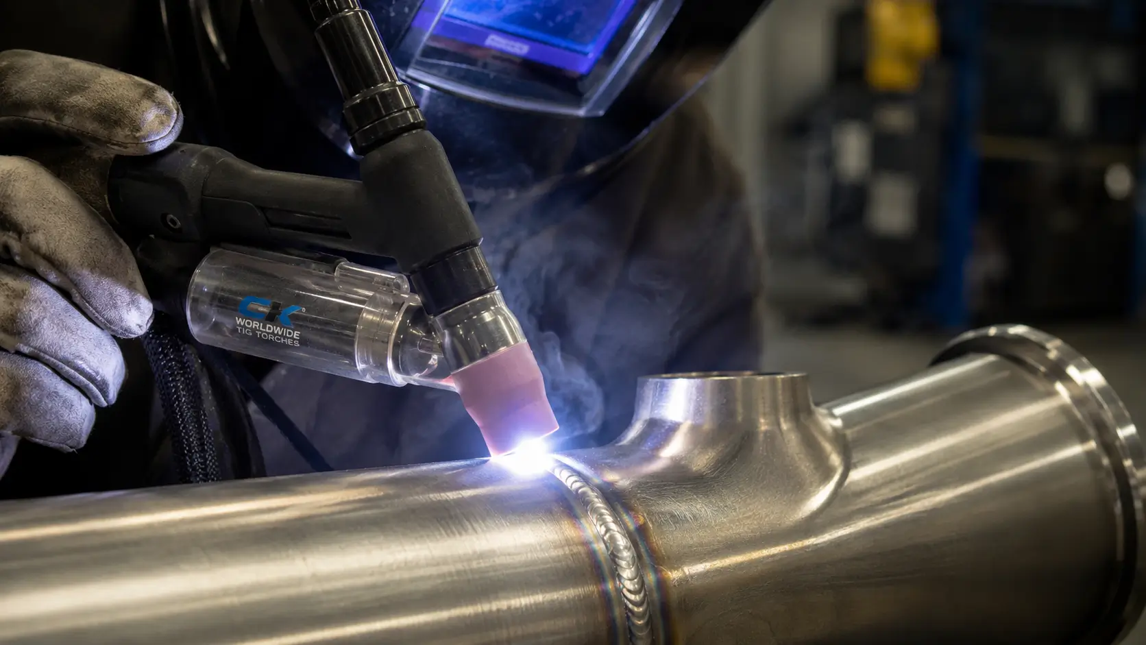 A close-up photograph showing a welder wearing a welding helmet and heavy gloves, operating a 'CK WORLDWIDE TIG TORCHES' brand TIG torch with a clear gas cup. A bright blue-white arc is produced at the tip of the torch, performing a precise TIG weld on a stainless steel pipe T-joint. The completed weld bead is neat and features rainbow-like heat tinting. Smoke rises from the weld point against a blurred industrial workshop background.