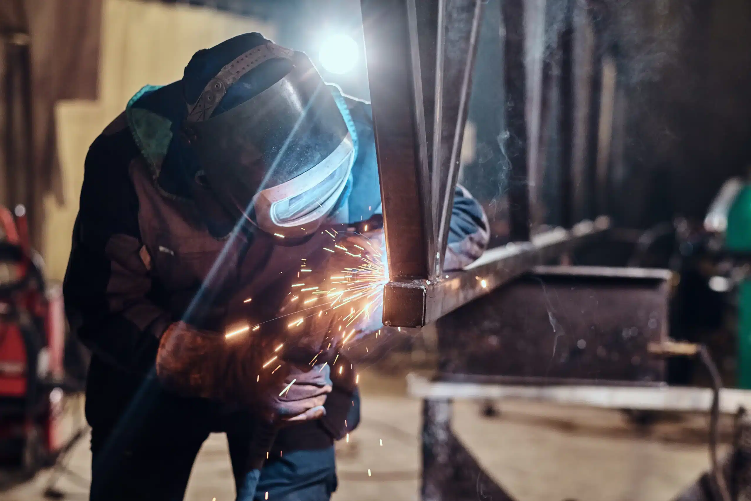 Worker welding a metal frame in a factory workshop with sparks flying during steel fabrication.