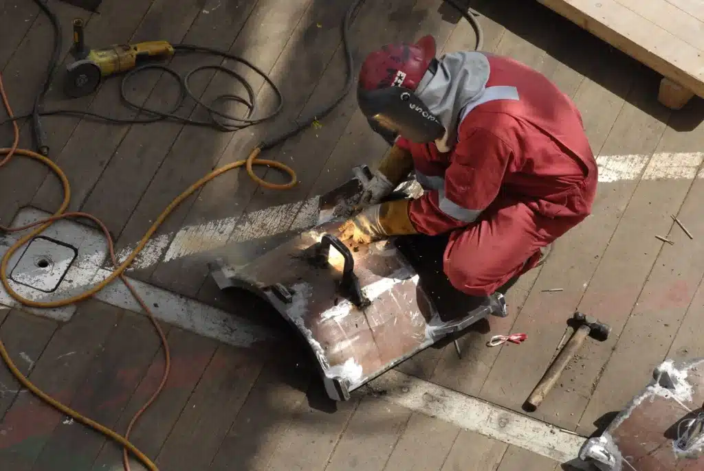 Welder wearing protective gear working on a thick metal part in an industrial fabrication area.